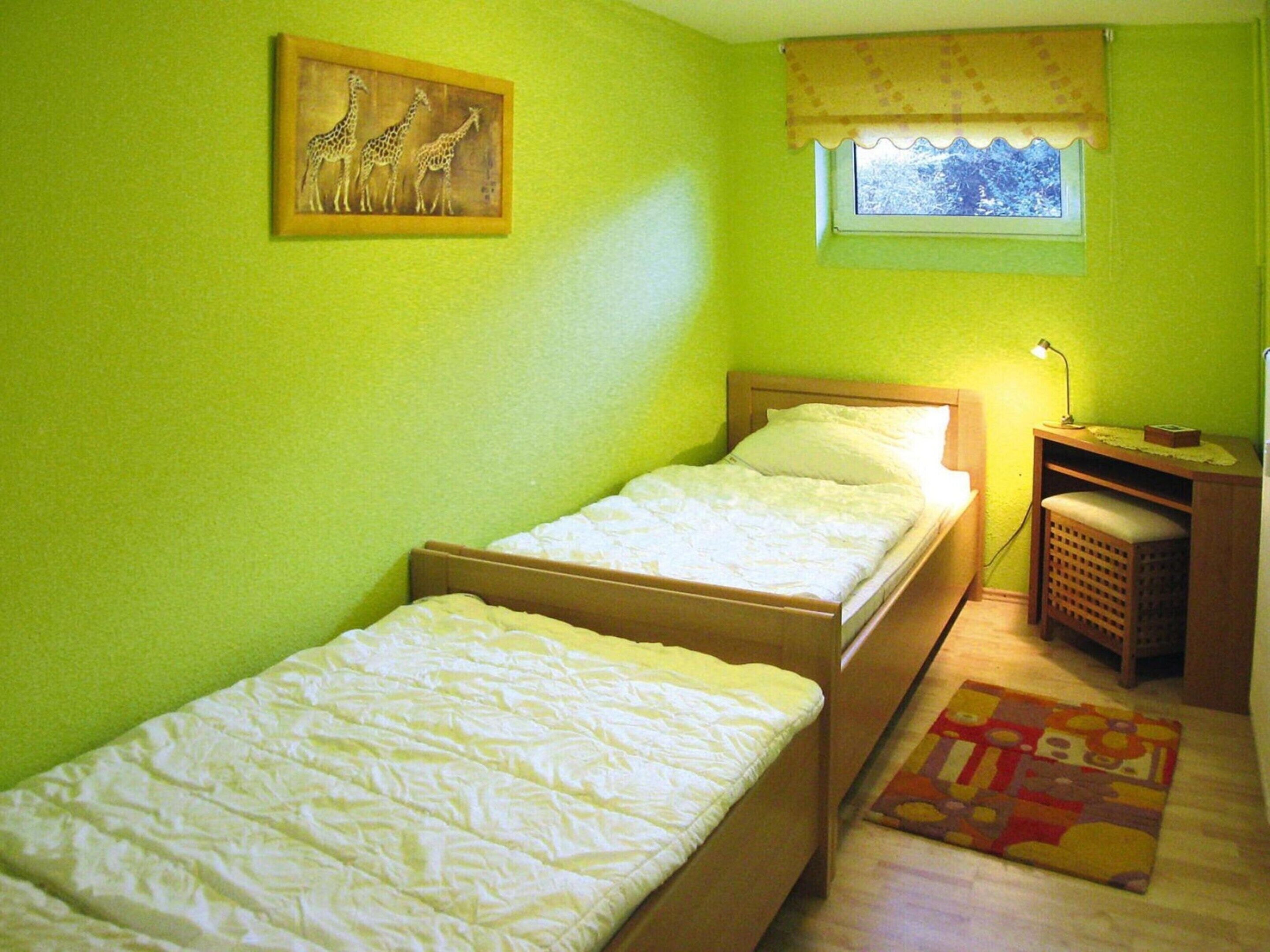 Quarto pequeno com uma cama de solteiro, cabeceira de madeira, mesa de cabeceira com luminária, pequena mesa com luminária e janela com vista para árvores.