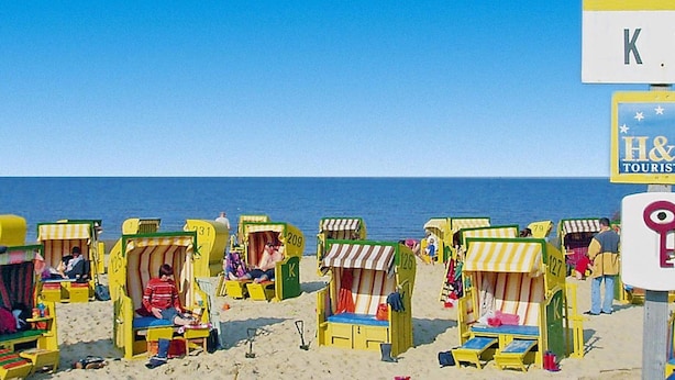 A beach with colorful beach chairs and umbrellas, a clear blue sky, and a sign for H& T TOURIS. - Ferienwohnung in Cuxhaven-duhnen (Cuxhaven)