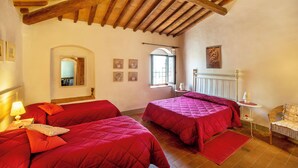 A room with two beds, wooden beams on the ceiling, a window with a view, and a small table with a lamp.