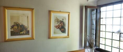 Une pièce avec deux reproductions botaniques encadrées accrochées au mur, une fenêtre donnant sur un paysage et un vase contenant des fleurs séchées posé sur une table en bois.