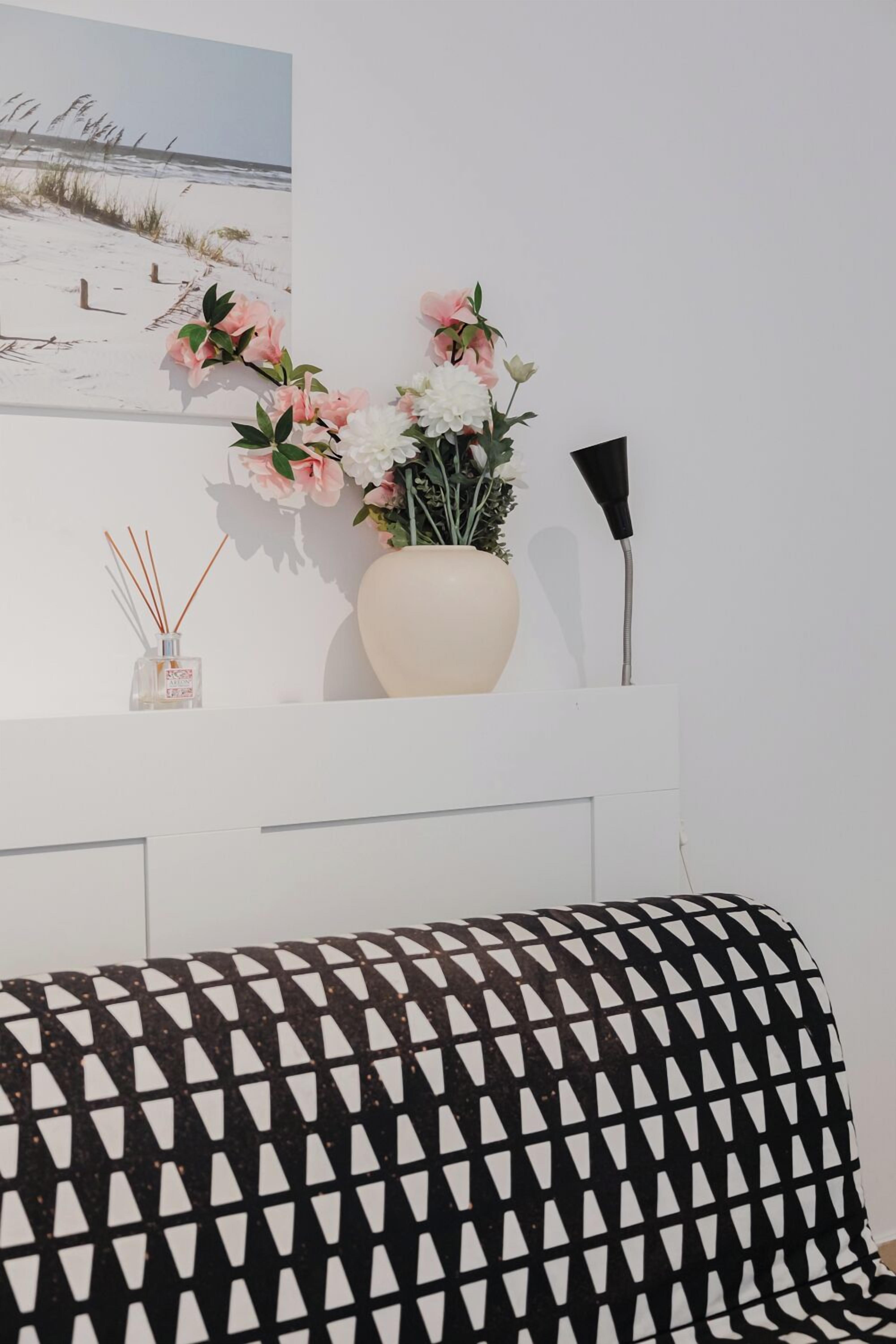 A white wall with a framed beach photograph, a vase with pink and white flowers, and a black and white patterned cushion.