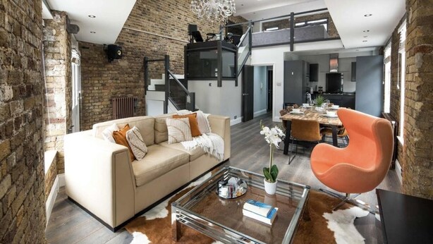 Un loft spacieux avec de hauts plafonds, des murs en briques apparentes, une cuisine moderne et un coin repas agrémenté d’une table basse en verre et d’une chaise orange.
