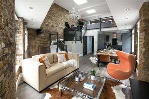Un loft spacieux avec de hauts plafonds, des murs en briques apparentes, une cuisine moderne et un coin repas agrémenté d’une table basse en verre et d’une chaise orange.