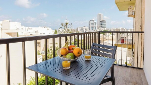 A balcony with a table set for two, a bowl of fruit, and a glass of orange juice.