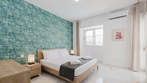 A modern hotel room with a large bed, a wooden nightstand, a desk, a window with a view, and a teal textured wall.