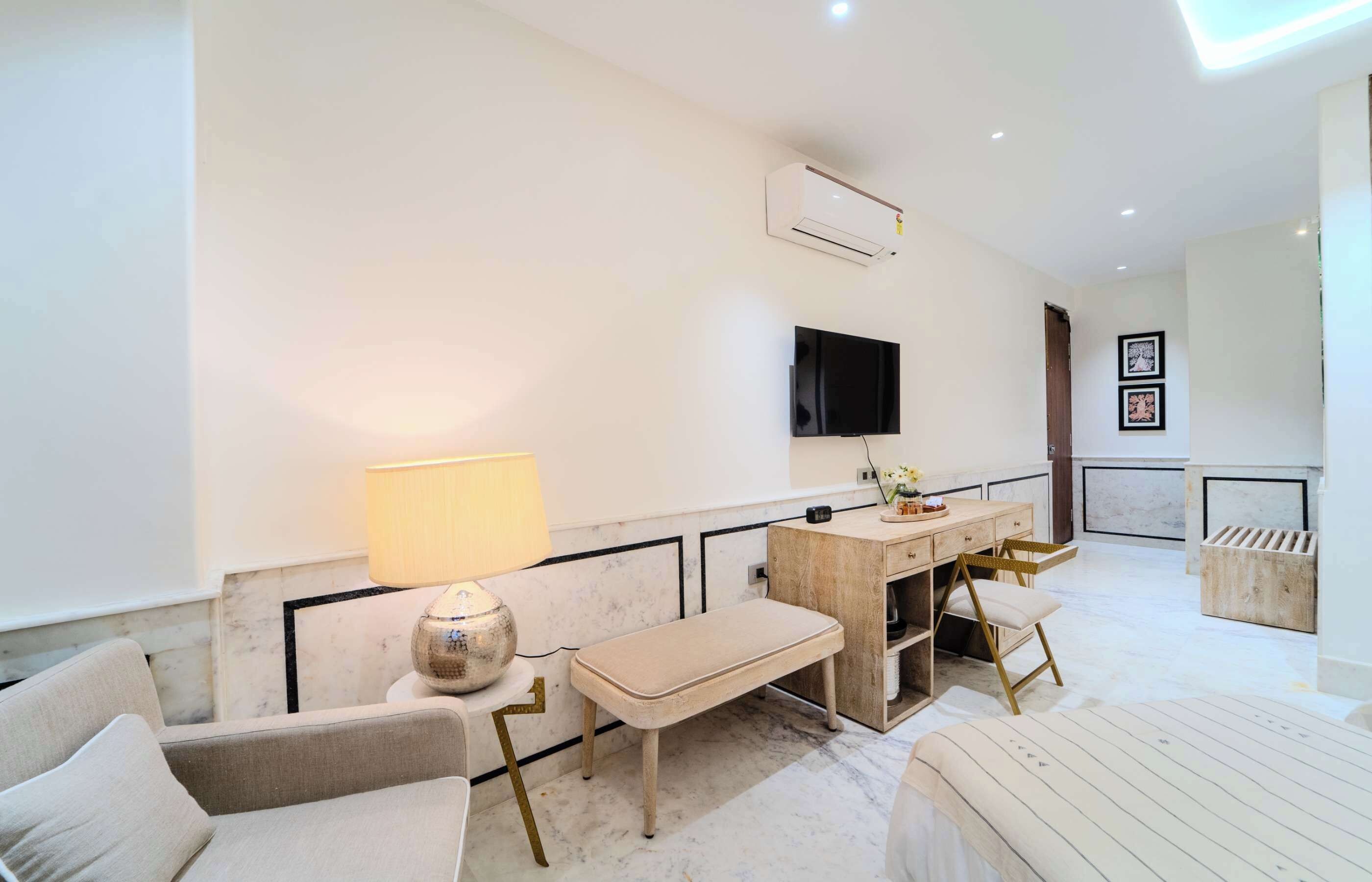 A modern hotel room with a sofa, a bench, a desk with a chair, a TV mounted on the wall, and an air conditioning unit.