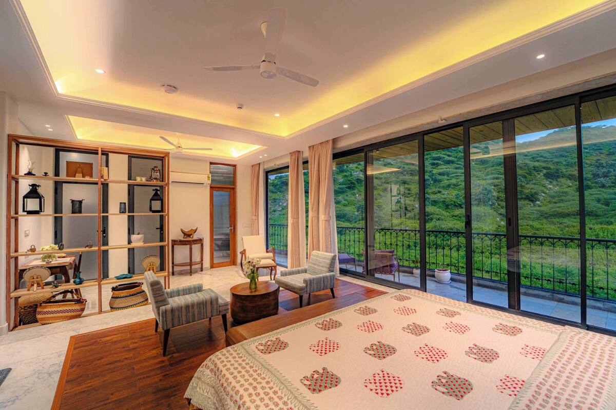 A spacious bedroom with a large bed, a seating area with chairs, a wooden floor, a built-in bookshelf, and a balcony with a scenic view.