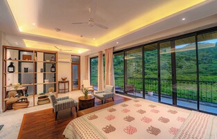 Un dormitorio espacioso con una cama grande, una zona de estar con sillas, suelo de madera, estantería empotrada y un balcón con vistas panorámicas.