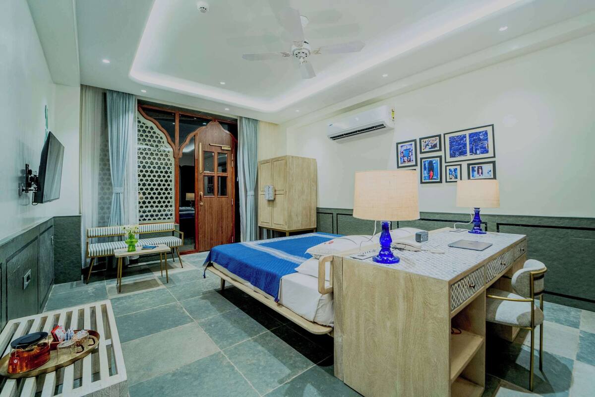 A hotel room with a bed, a desk with a lamp, a chair, a wardrobe, a TV, and a wall with framed pictures.