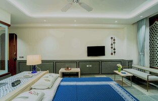 Habitación de hotel con cama, mesita de noche, lámpara, televisor y zona de estar con un banco.