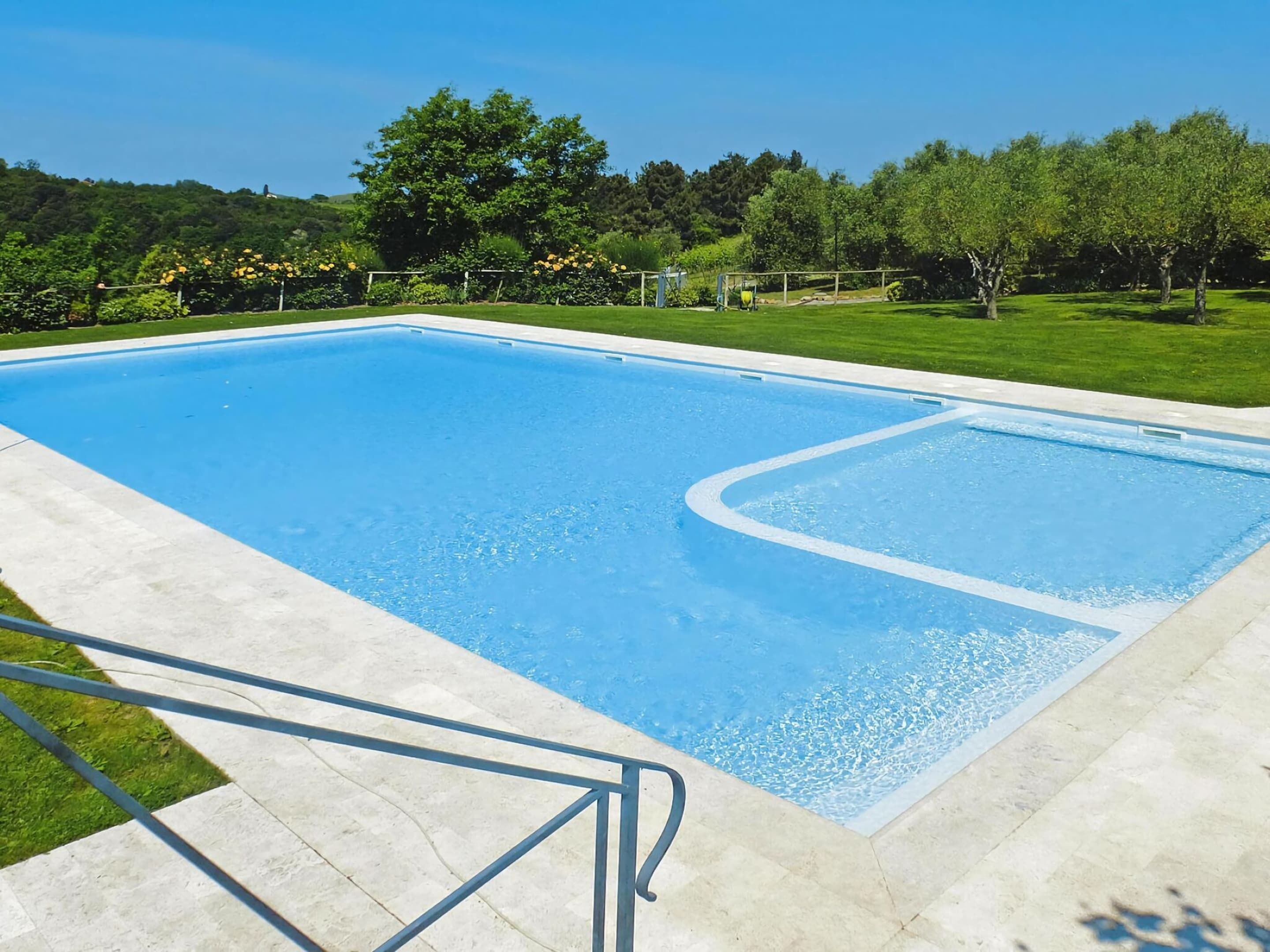 A rectangular swimming pool with a clear blue water surface, surrounded by a concrete deck and a grassy area with trees.