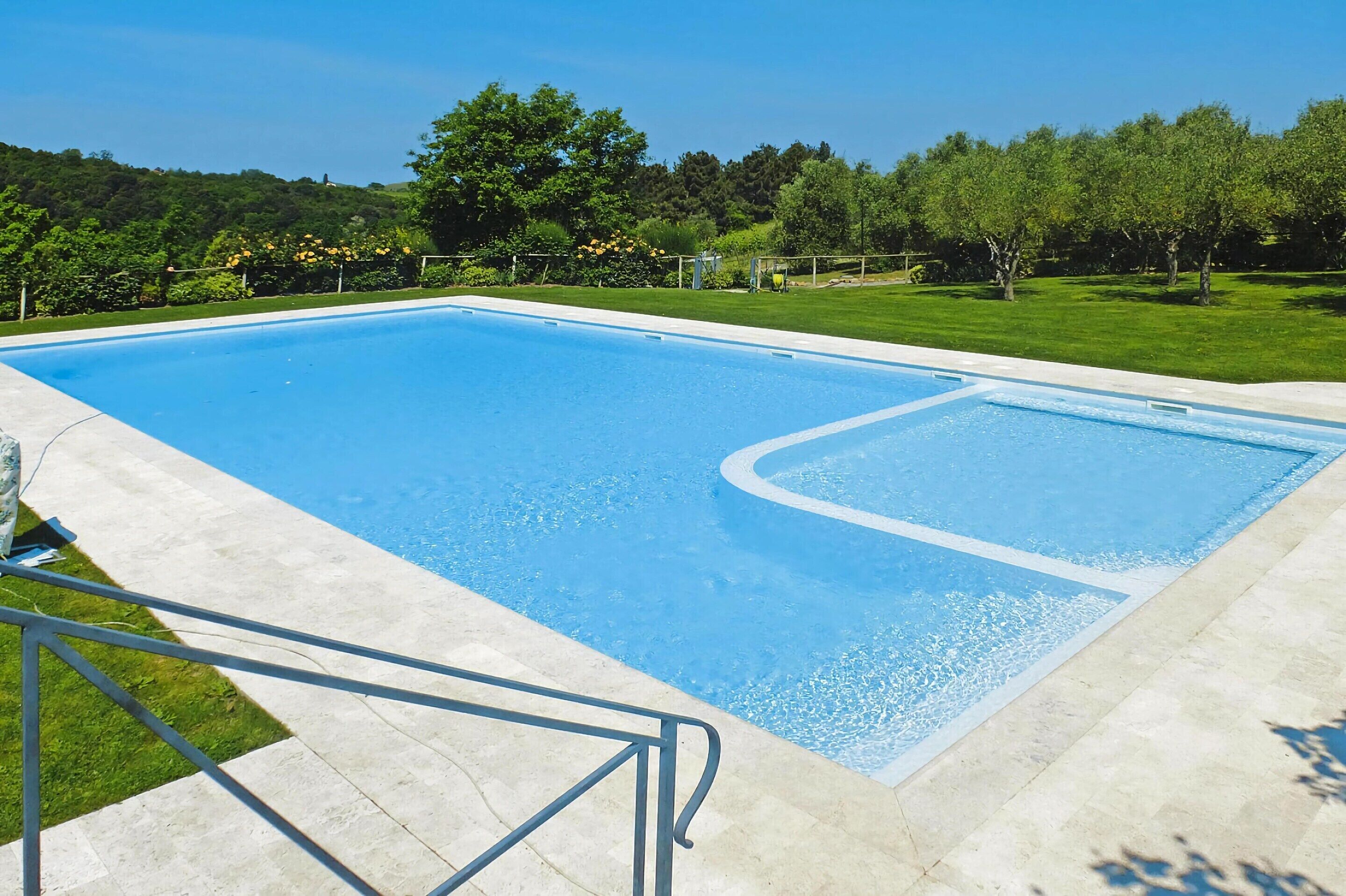 Piscina retangular com superfície de água azul cristalina, rodeada por um deck de concreto e uma área gramada com árvores.