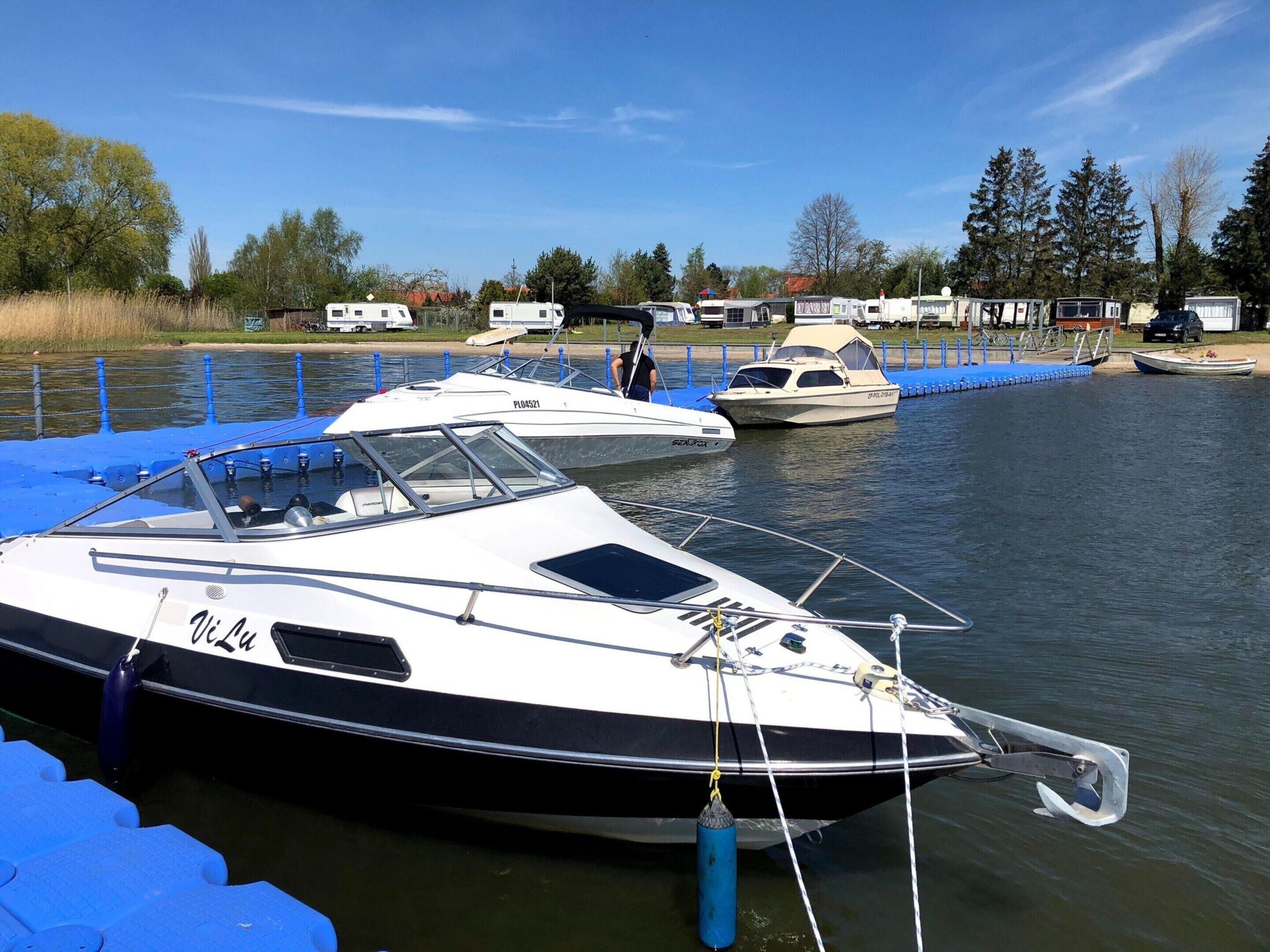 A marina with several boats docked, a blue floating dock, and a row of houses in the background.