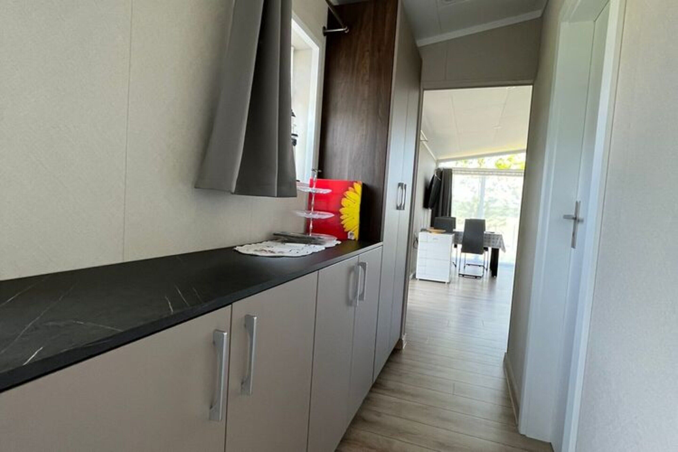 Un couloir étroit mène à une cuisine avec un plan de travail noir et des placards gris. Un objet décoratif rouge et jaune est posé sur le plan de travail. Le couloir donne sur un coin repas avec une table et des chaises.
