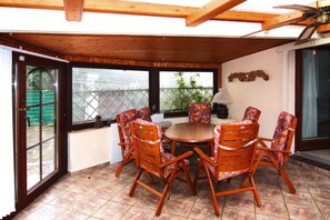 A covered patio with a round wooden table and six chairs, large windows, and a ceiling fan. - Holiday Home in Busum in a Beautiful Area (Büsum)