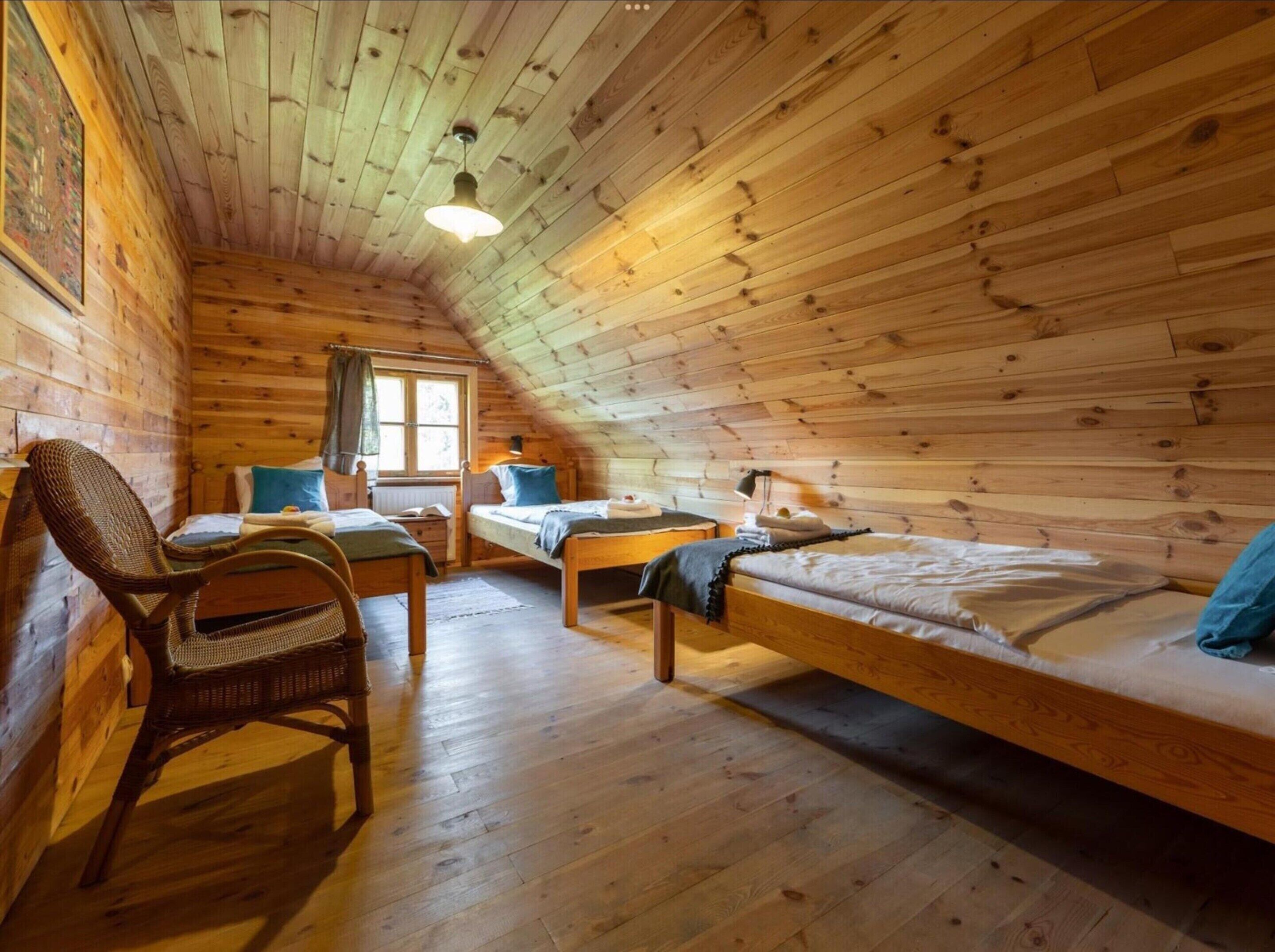 A cozy cabin with wooden walls and floors, two single beds, a wicker chair, and a small table.