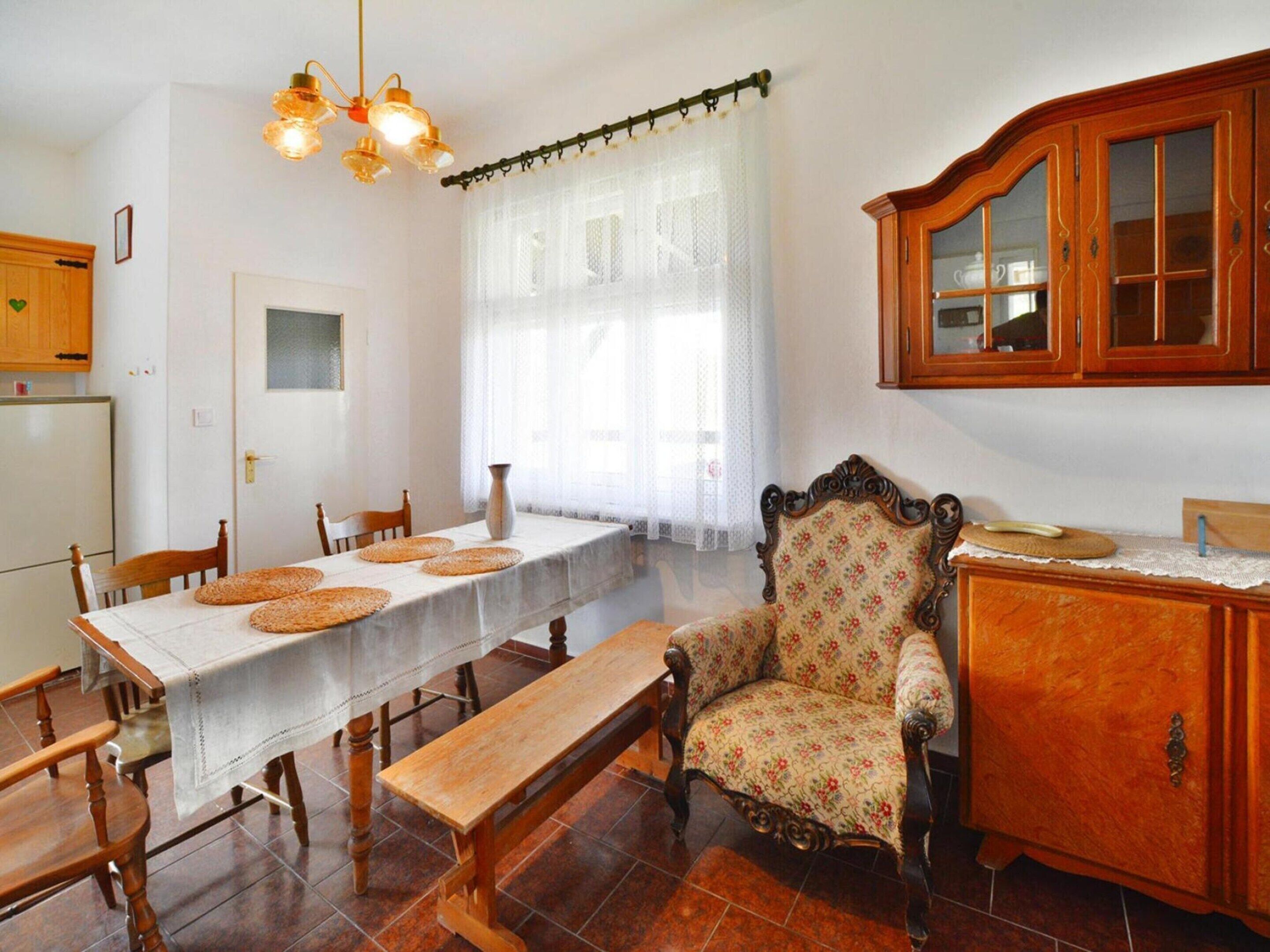 Un comedor con una mesa de madera, sillas, un banco, un sillón con estampado floral, un armario de madera y una araña.