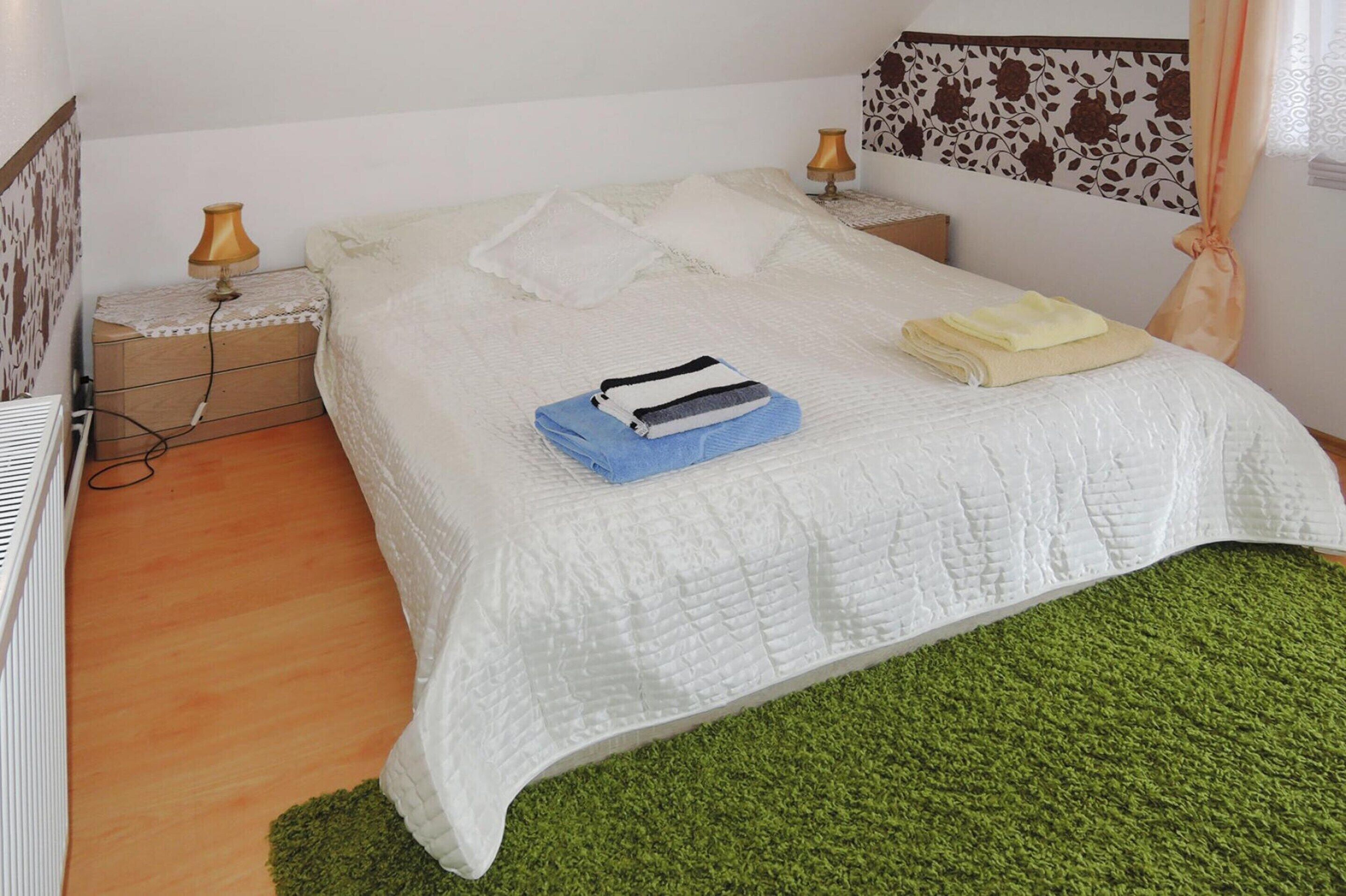 Ein ordentlich bezogenes Bett mit weißer Steudecke, zwei Kissen und gefalteten Handtüchern. Ein Nachttisch mit Lampe und Steckdose ist vorhanden. Der Raum verfügt über einen grünen Teppich und einen blumenmusterigen Kopfteil.
