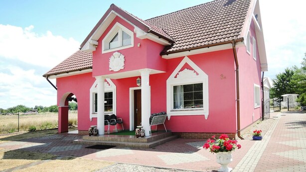 Una casa rosa con tetto in tegole, un portico con sedie e il numero '3A' sul lato.