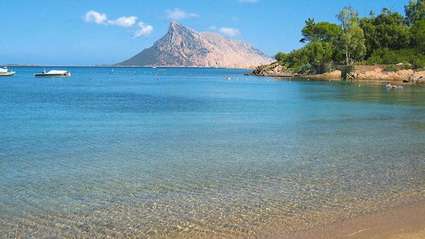 Una playa con agua cristalina, barcos y una montaña al fondo.