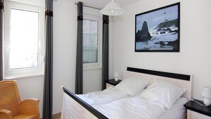 A neatly arranged bedroom with a bed, a chair, a nightstand, a lamp, and a painting of a lighthouse on the wall. - Ferienwohnung, Graal-muritz (Graal Mueritz)