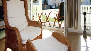 A wicker chair and ottoman in a sunlit room with a view of a residential area through large windows. - Ferienwohnung, Graal-muritz (Graal Mueritz)