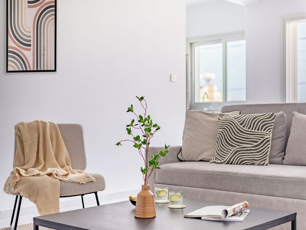 A modern living room with a sofa, a coffee table, a chair with a blanket, a vase with a plant, and a framed picture on the wall. - Sanders Cruise Limassol (Limassol)