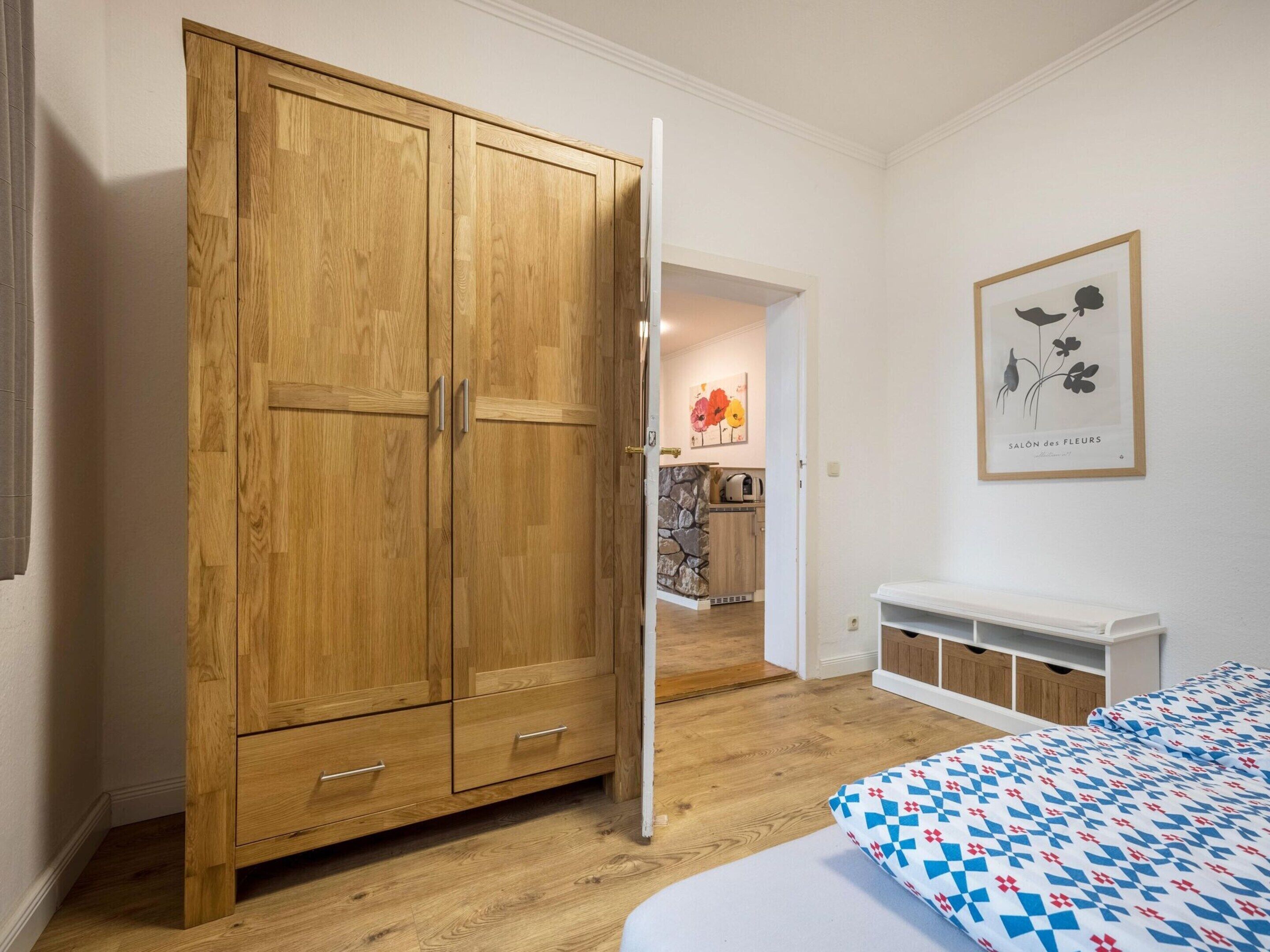 A bedroom with a wooden wardrobe, a bed with a patterned duvet, a white bedside table, and a framed picture on the wall.