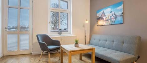 A room with a grey sofa, a wooden table, a grey chair, and two windows with shutters.