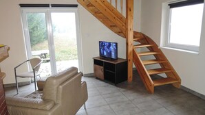 Un salon avec un escalier en bois, un téléviseur posé sur un meuble, un canapé et une porte-fenêtre coulissante donnant sur un espace extérieur.