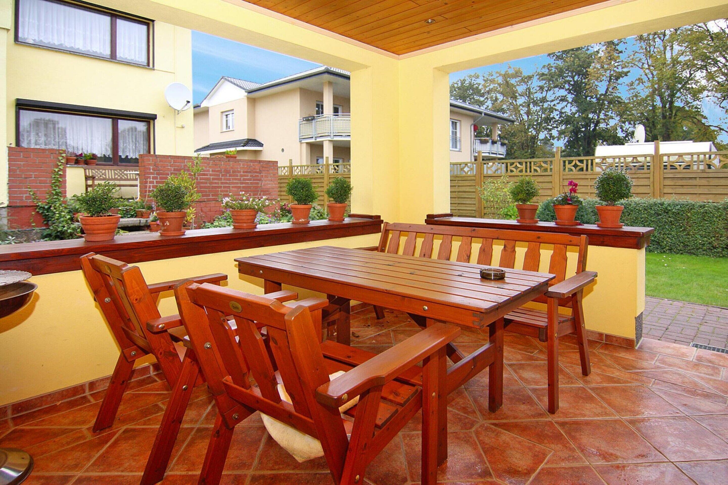 Une terrasse avec une table et des chaises en bois, entourée de plantes en pot et d’un mur en briques.