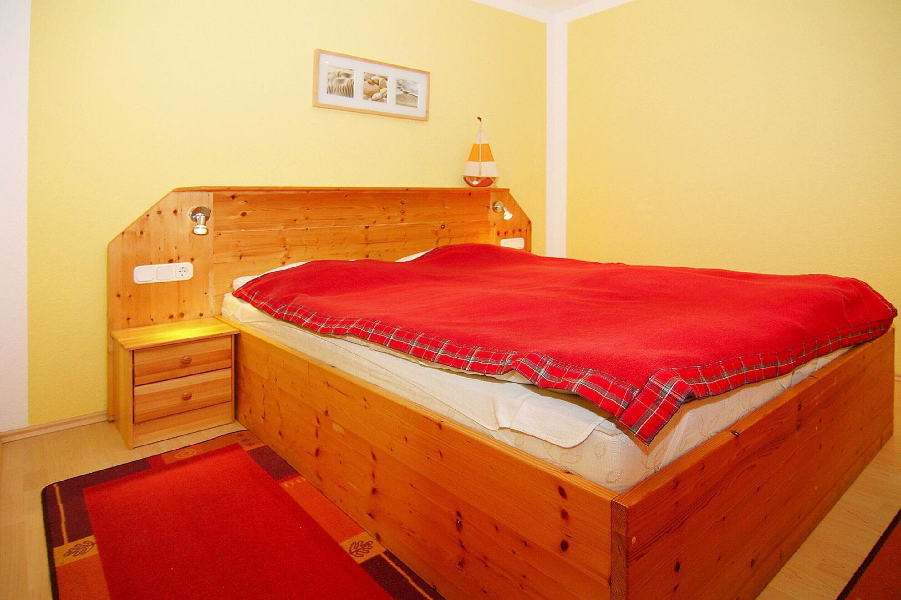 Ein hölzernes Bett mit roter Decke, ein Nachttisch und eine kleine Lampe.