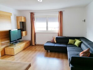 A living room with a television on a stand, a sofa, and a window with curtains. - Holiday on an Organic Farm in Hart im Zillertal (Hart im Zillertal)