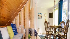 A cozy living room with a wooden staircase, a sofa with patterned cushions, a dining table with wicker chairs, and a view of the outdoors through a window. - Apartment in Jaroslawiec Near the Beach (Postomino)