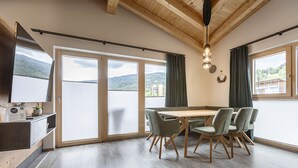 Un comedor moderno con una mesa y sillas de madera, ventanas amplias con cortinas, y vistas a un lago y montañas.