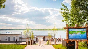 A pathway leading to a lakeside area with a wooden signboard, a small building, and a view of the lake. - Camp David Sport Resort (Rackwitz)
