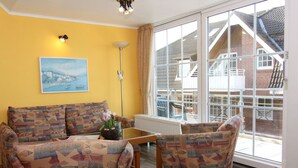 Ein Wohnzimmer mit gemusterten Sofas, einem Couchtisch und einer Stehlampe. Ein großes Fenster bietet Blick auf ein Backsteingebäude und eine Treppe.