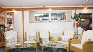Ein gemütliches Zimmer mit Korbstühlen, einem Glastisch, einem Fenster mit Segelbooten und einem Bücherregal mit Büchern und Pflanzen.