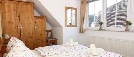 Un dormitorio con techo inclinado, armario de madera, dos camas individuales con ropa de cama estampada, una silla y una ventana con cortinas.