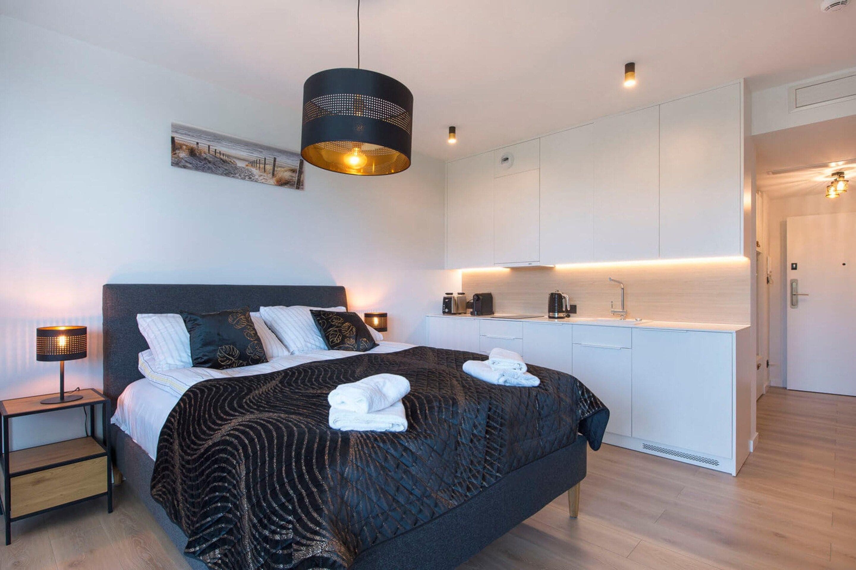 Un dormitorio moderno con una cama grande, lámparas de noche, un candelabro colgante, un cuadro y una pequeña cocina.