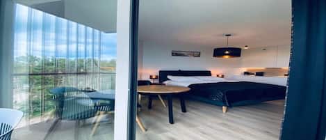 A modern hotel room with a large sliding glass door leading to a balcony, a bed with a dark headboard, a small round table, and a single chair.