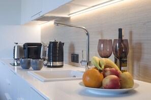 Eine moderne Küche mit weißer Arbeitsplatte, einer Kaffeemaschine, einem Wasserkocher, einer Obstschale mit Bananen und Äpfeln sowie zwei Weingläsern.