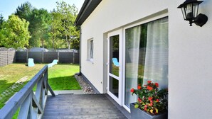 Una terraza de madera que conduce a una casa con puerta corrediza de vidrio, una ventana pequeña y un accesorio de luz exterior fijado a la pared.