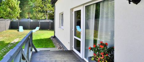 Una terraza de madera que conduce a una casa con puerta corrediza de vidrio, una ventana pequeña y un accesorio de luz exterior fijado a la pared.