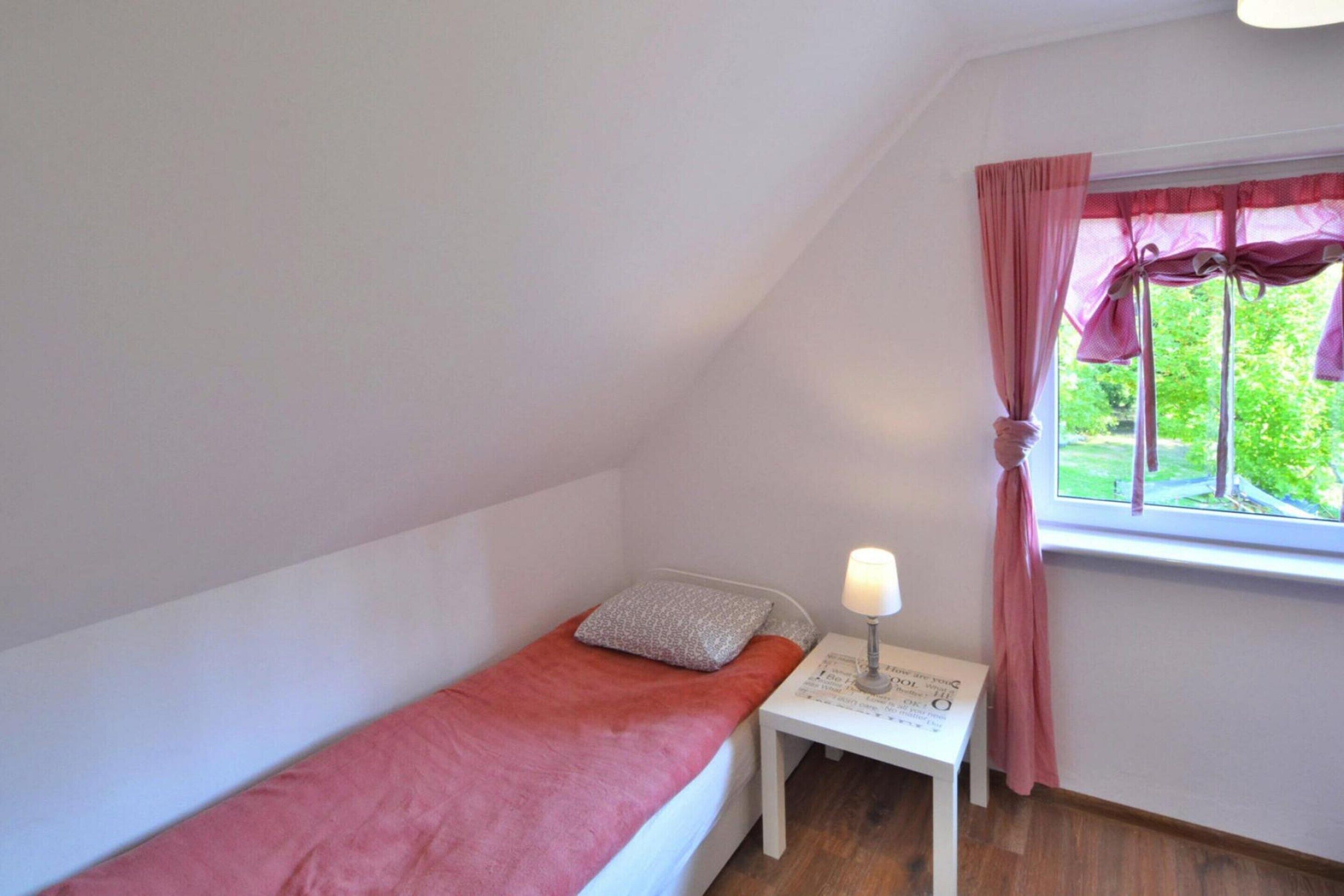 Quarto pequeno com uma cama de solteiro, janela, mesa de cabeceira com abajur e cortina rosa.