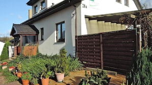 A two-story house with a white exterior and a brown roof. There is a garage with a brown door and a small porch with a tiled roof. The house is surrounded by various potted plants and shrubs. - Agriculture - Studios, Borzyslawiec by Lubczyna (Goleniow)