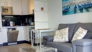 Ein modernes Wohnzimmer mit grauem Sofa, einem Glastisch und einer Küchenzeile mit weißen Schränken und einer Mikrowelle.