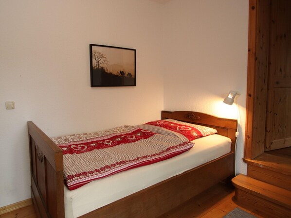 Una cama individual con cabecera de madera, una colcha estampada en rojo y blanco, y un cuadro colgado en la pared.