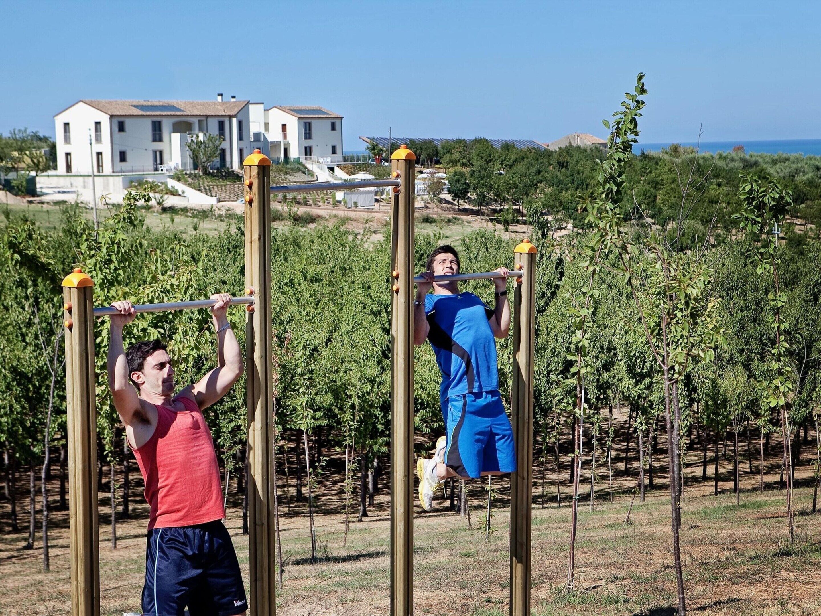 Two people exercising on outdoor gym equipment with a vineyard and buildings in the background.