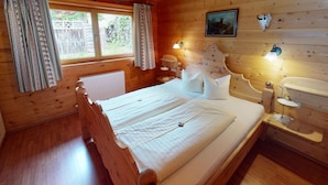 Quarto de cabana de madeira com uma cama, duas luminárias de cabeceira, um radiador e uma janela com cortinas.